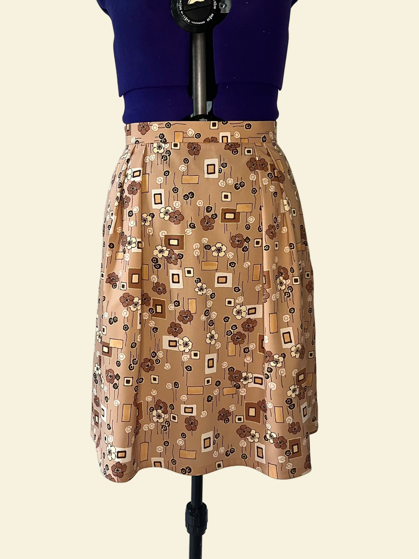 Mannequin wearing a skirt. The skirt has four pleats on the front and four pleats on the back, and inseam pockets. The fabric features brown/beige tones and geometric shapes and flowers.