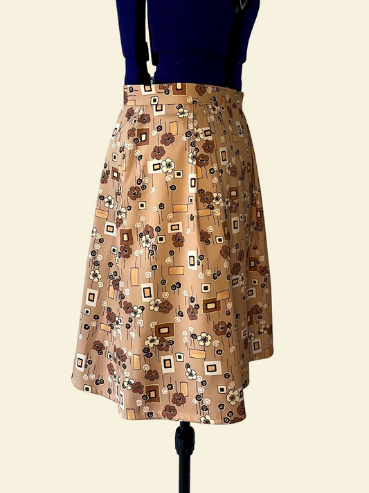 Mannequin wearing a skirt. The skirt has four pleats on the front and four pleats on the back, and inseam pockets. The fabric features brown/beige tones and geometric shapes and flowers.